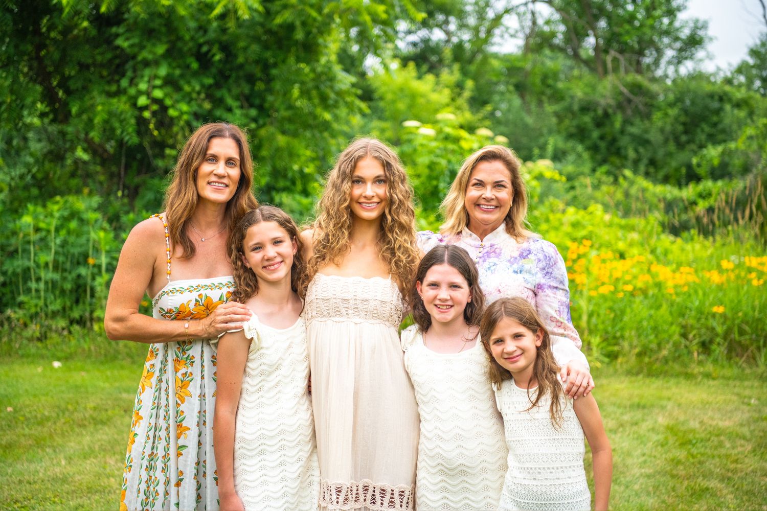 A group in light-colored dresses pose together in a lush summer garden with yellow wildflowers in the background.