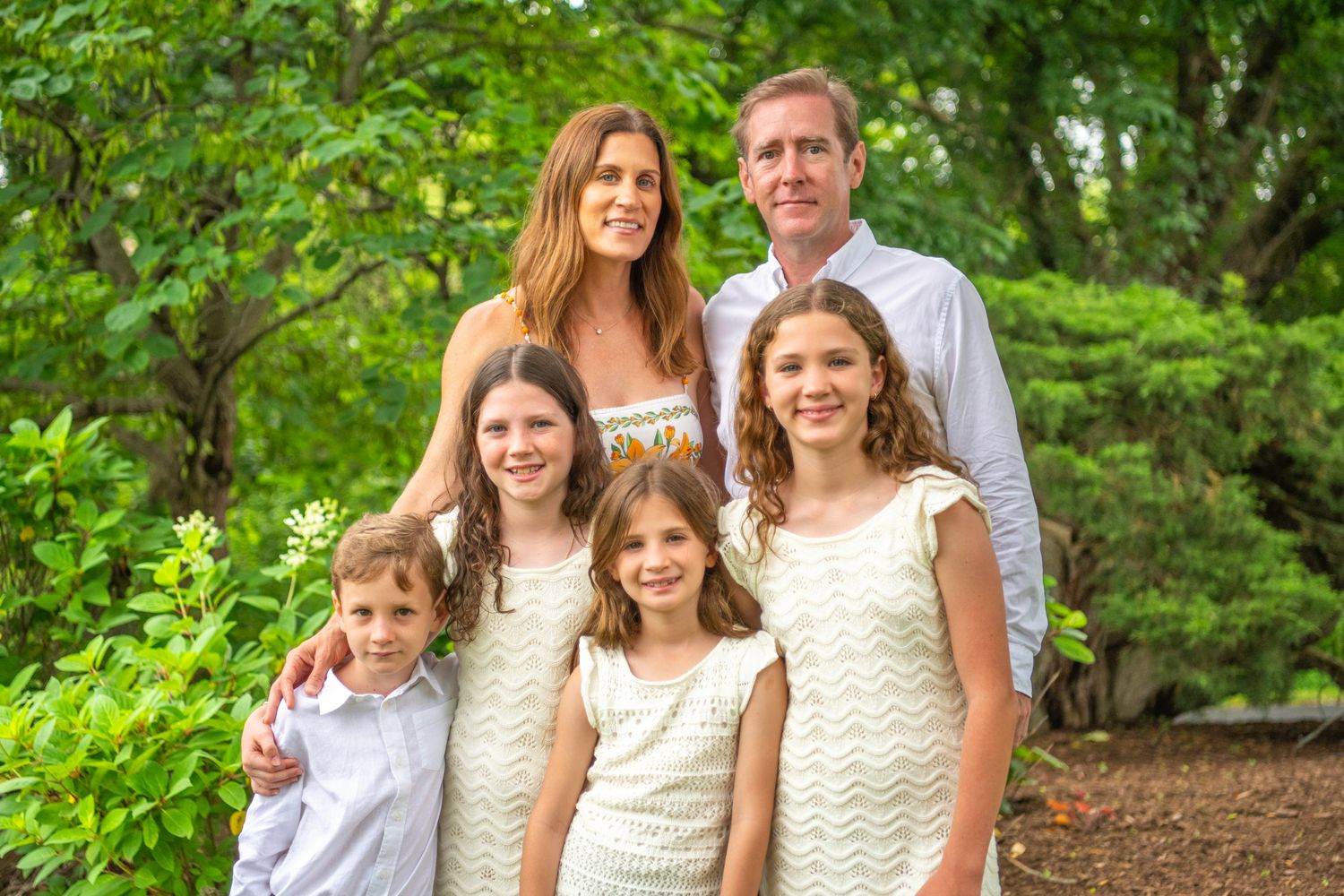 Multigenerational family group poses outdoors in casual attire surrounded by lush green foliage in summer setting.
