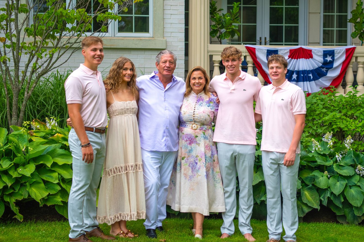 A group of people wearing white and pink outfits pose together in a garden setting against a white house.