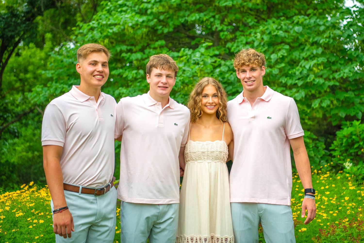 A group of four people in casual summer attire pose together outdoors against a lush green forest background.