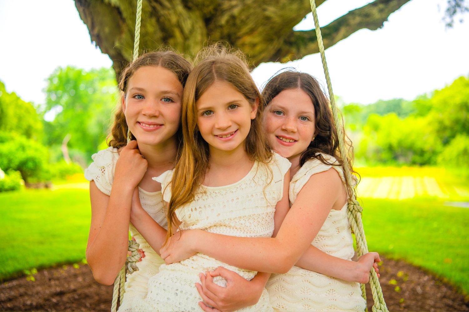 Three children in white dresses smile together while sitting on a swing in a lush green garden setting.