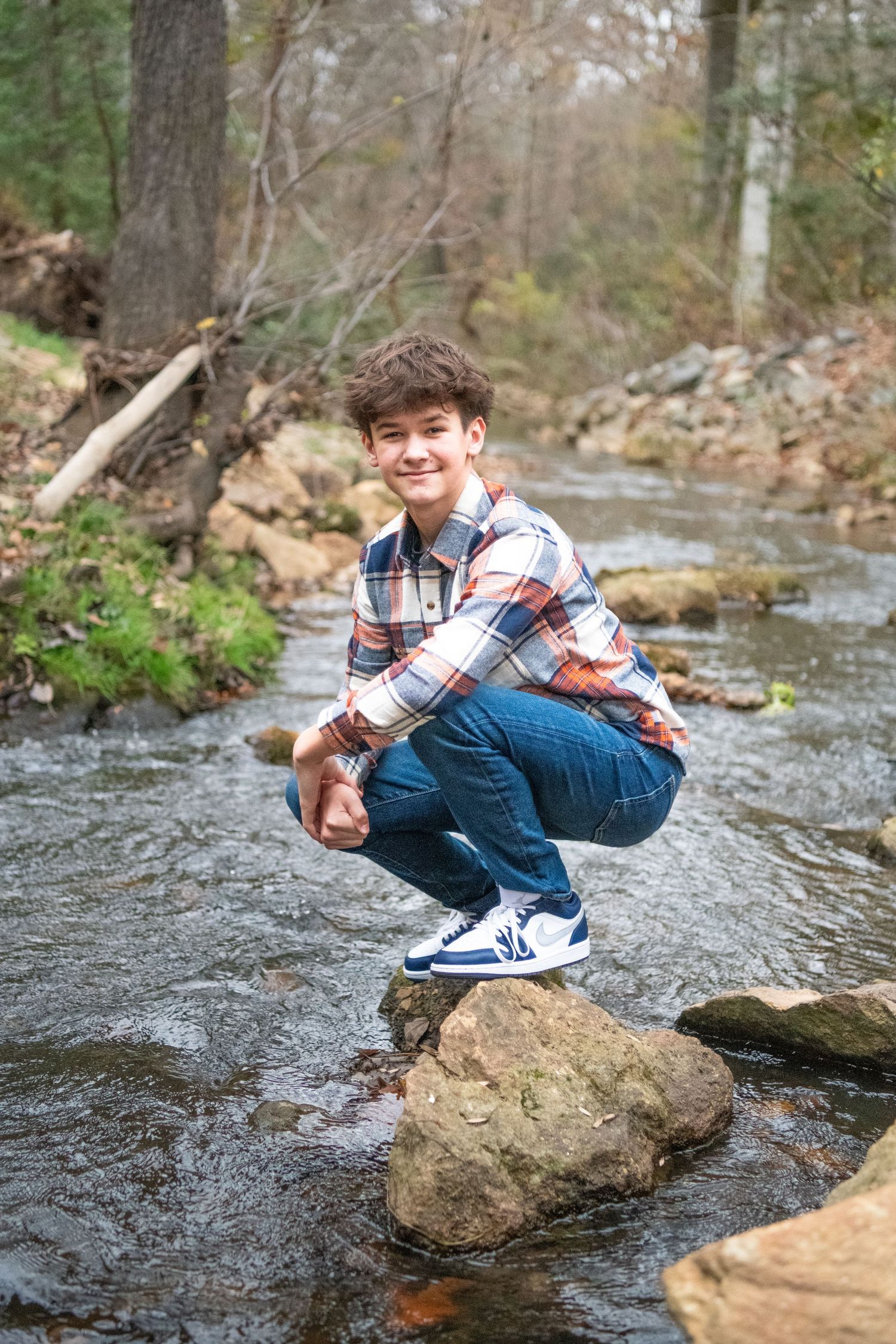 Person crouching on rocks in a flowing stream wearing plaid and jeans.