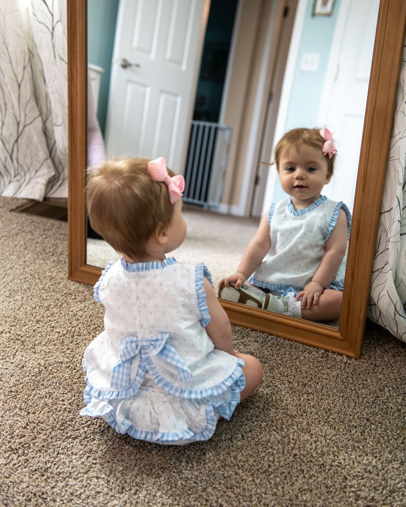 Small child in blue dress looks at reflection in full-length mirror.
