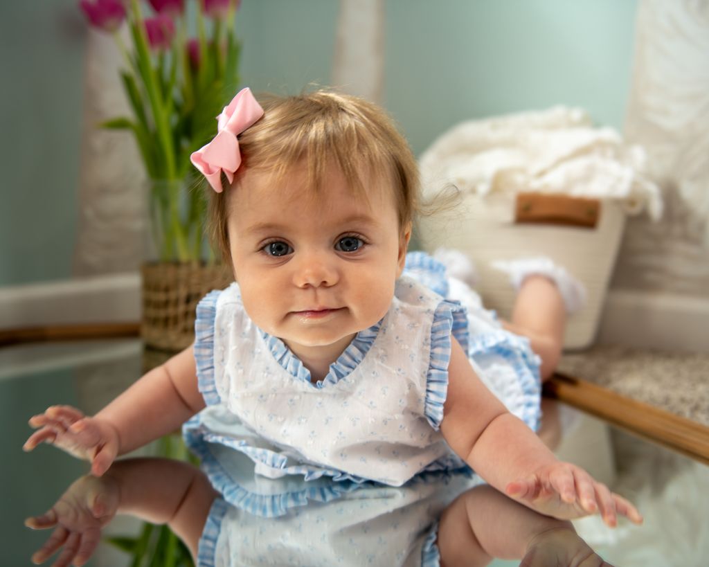 Baby wearing blue and white dress sitting indoors near potted tulips.