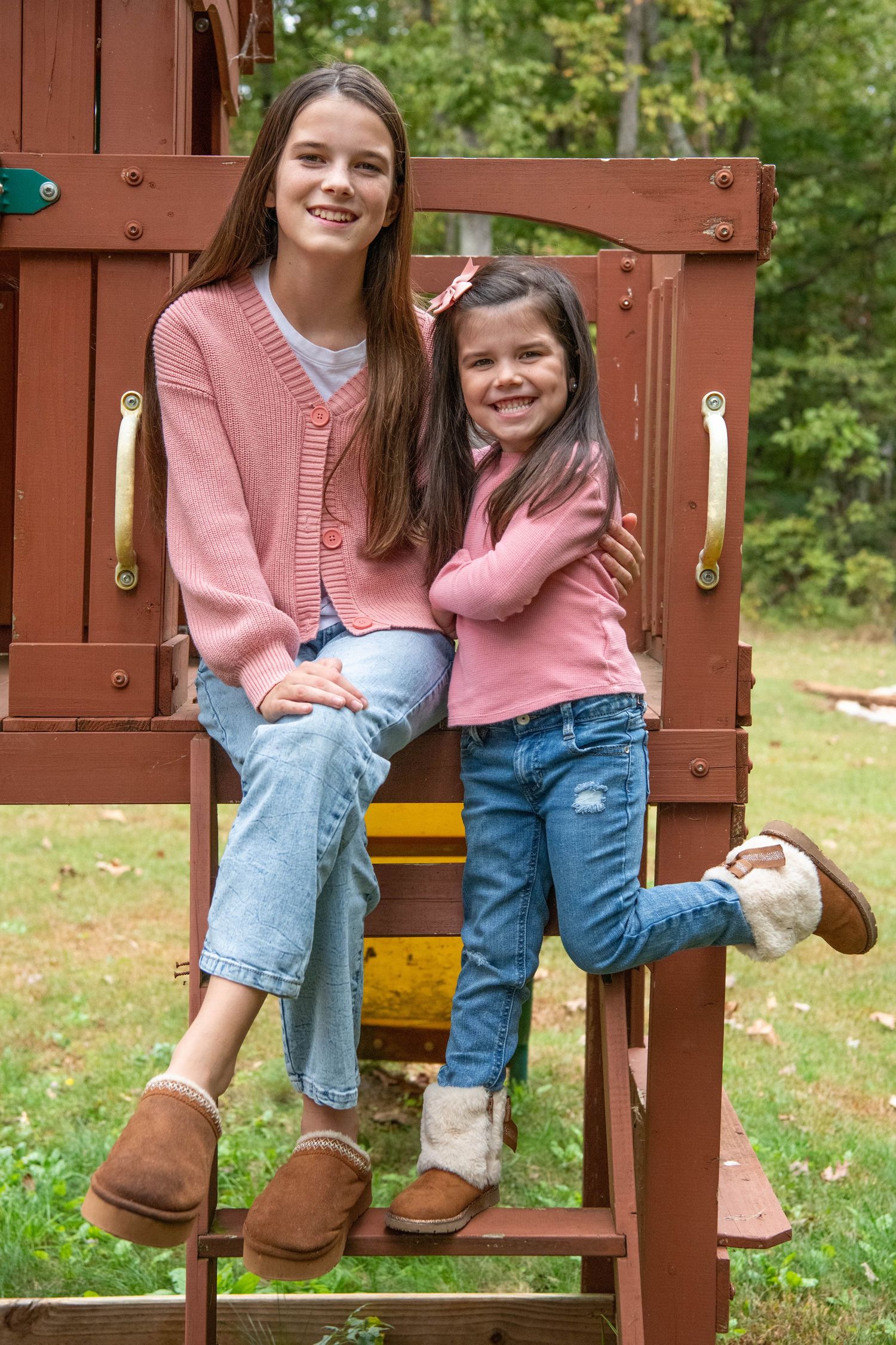 Two people in matching pink sweaters and jeans sit together on wooden playground equipment in an outdoor setting.