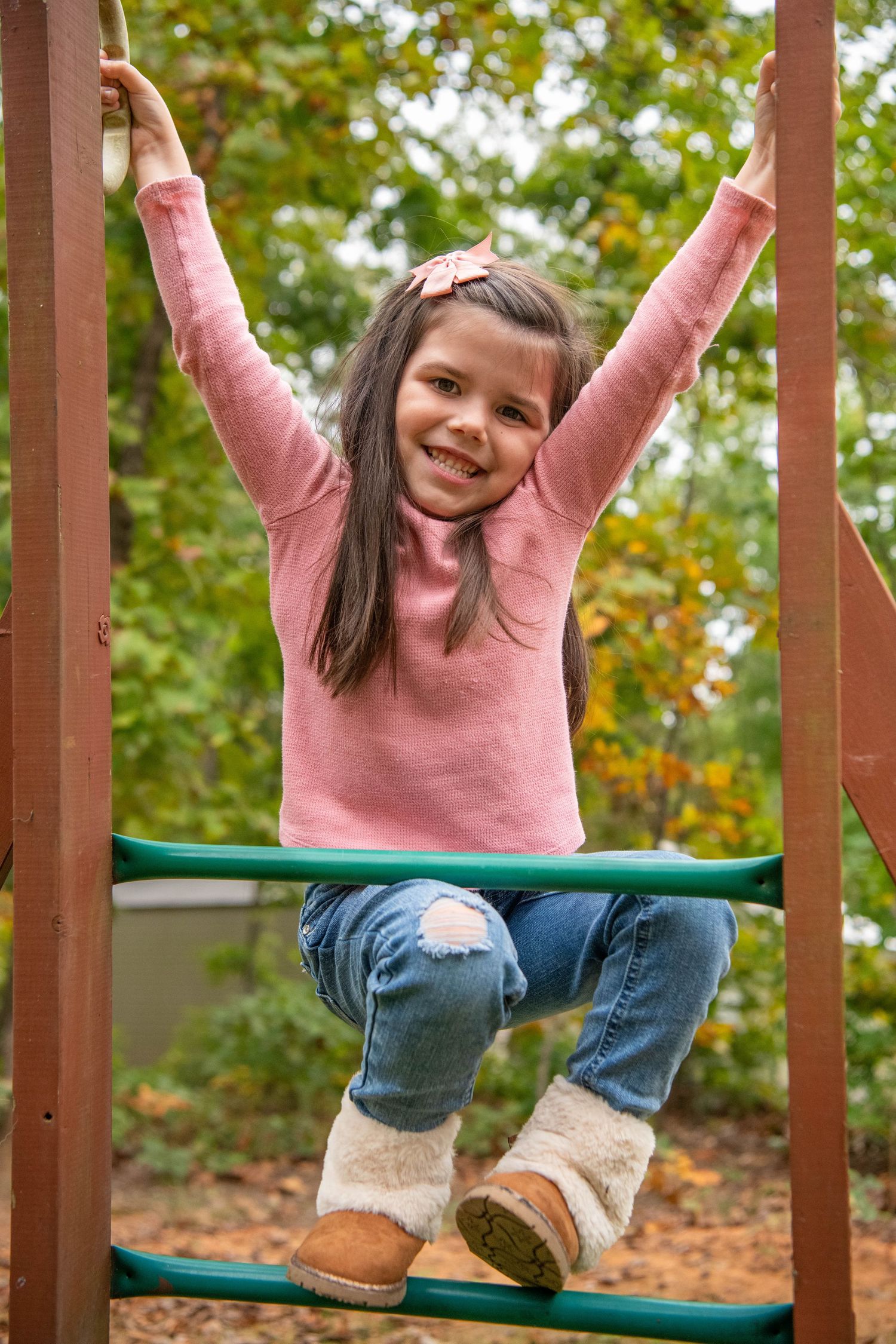 Someone wearing a pink sweater and jeans hangs upside down on playground equipment in an autumn setting.