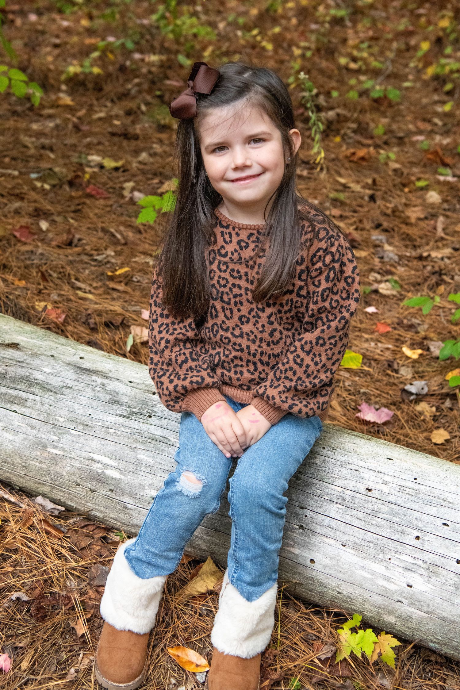 Someone in a leopard print sweater and jeans sits casually on a fallen log surrounded by autumn leaves.