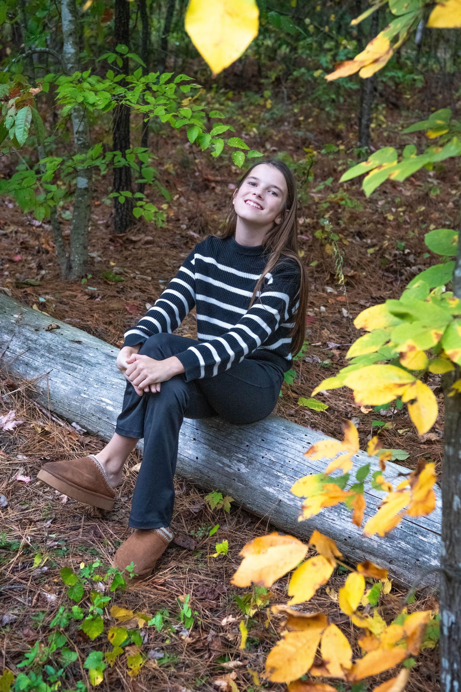 A person in a striped sweater sits on a fallen log surrounded by autumn leaves in a wooded setting.