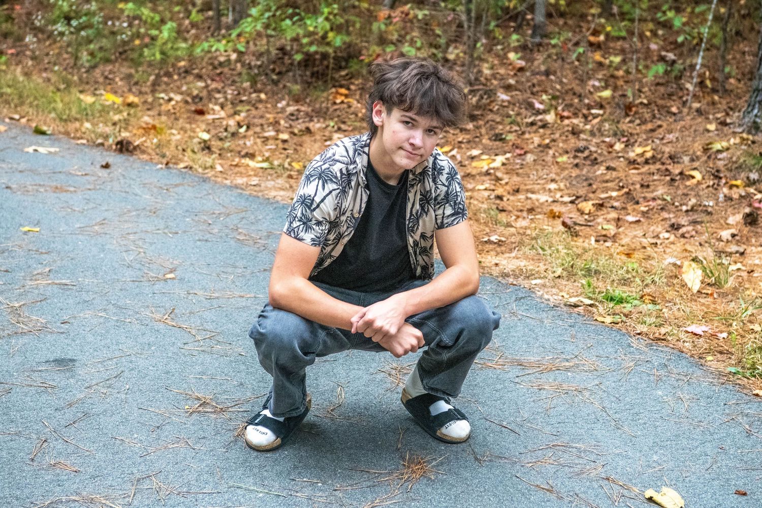 A person crouches down on a paved path surrounded by fallen autumn leaves in a park setting.