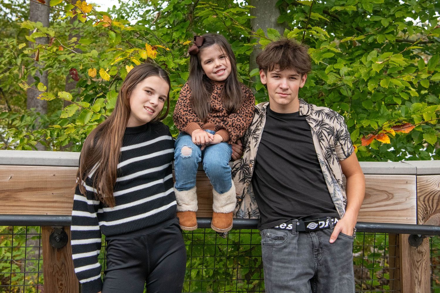 Three young people pose casually together on a wooden railing with autumn foliage in the background.