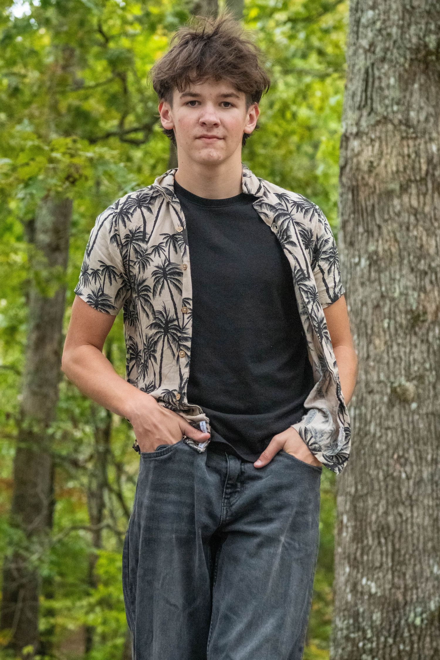 A person wearing a patterned shirt and dark jeans poses against tree trunks in a wooded setting.