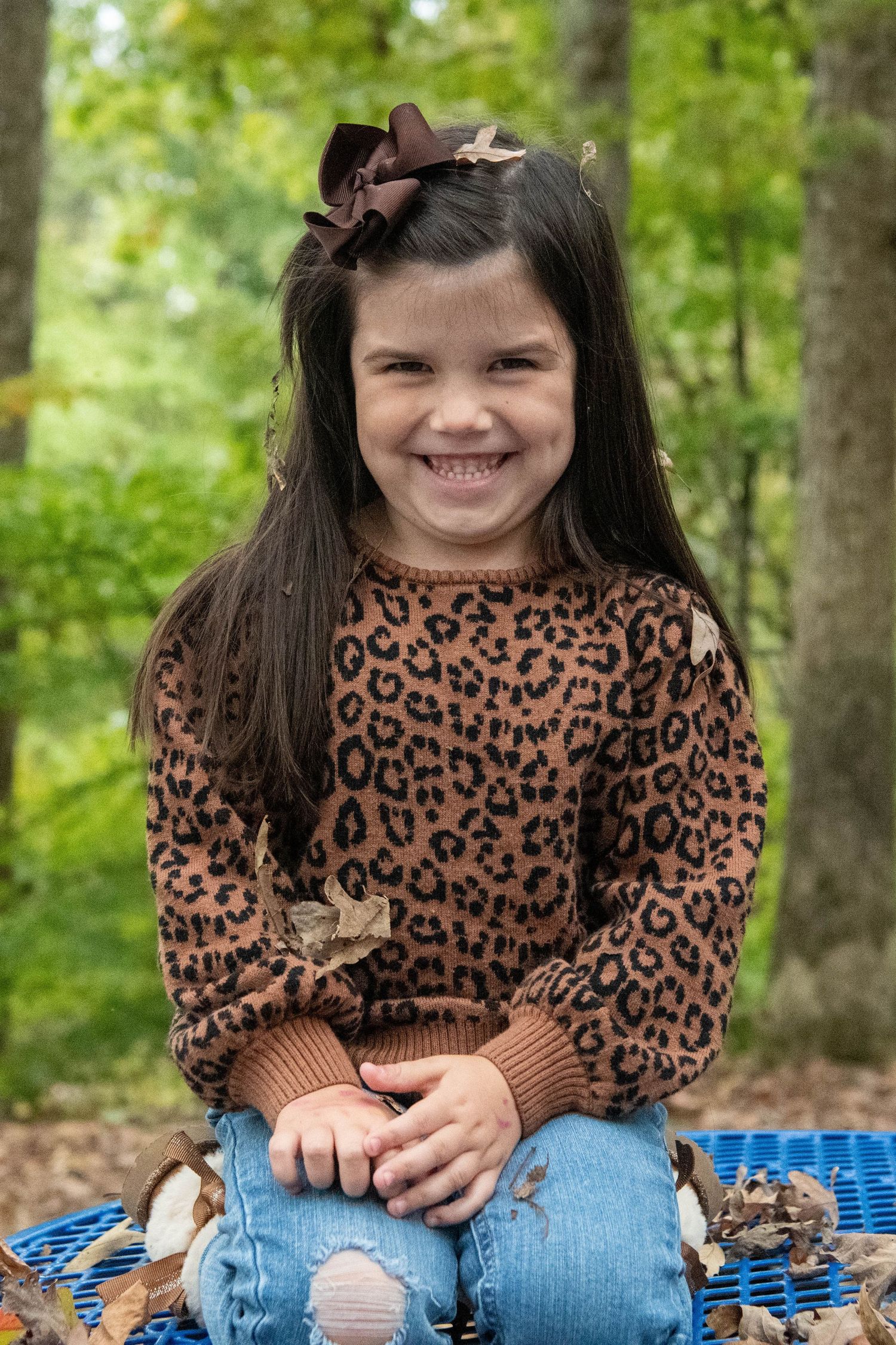 A child in a leopard print sweater sits on a blue blanket outdoors while smiling at the camera.
