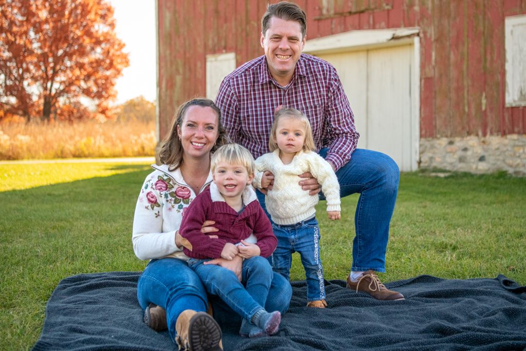A family of four sits together on a blanket in front of a red barn.