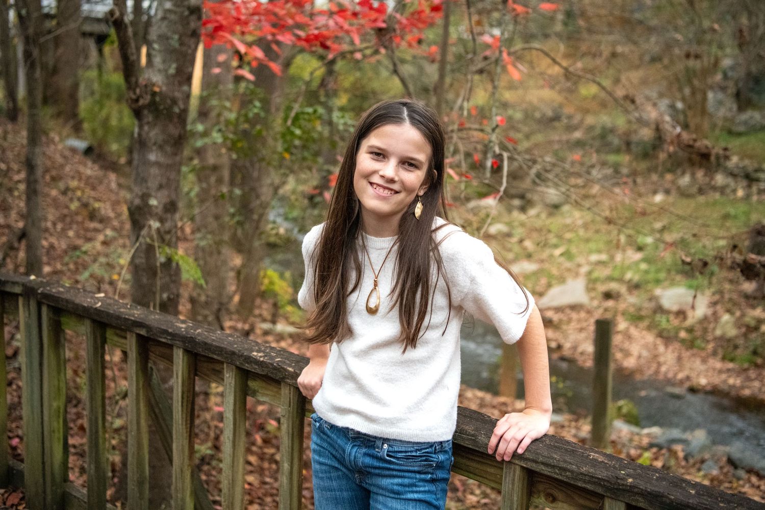 Person in white shirt and jeans standing by wooden railing with autumn foliage in background.