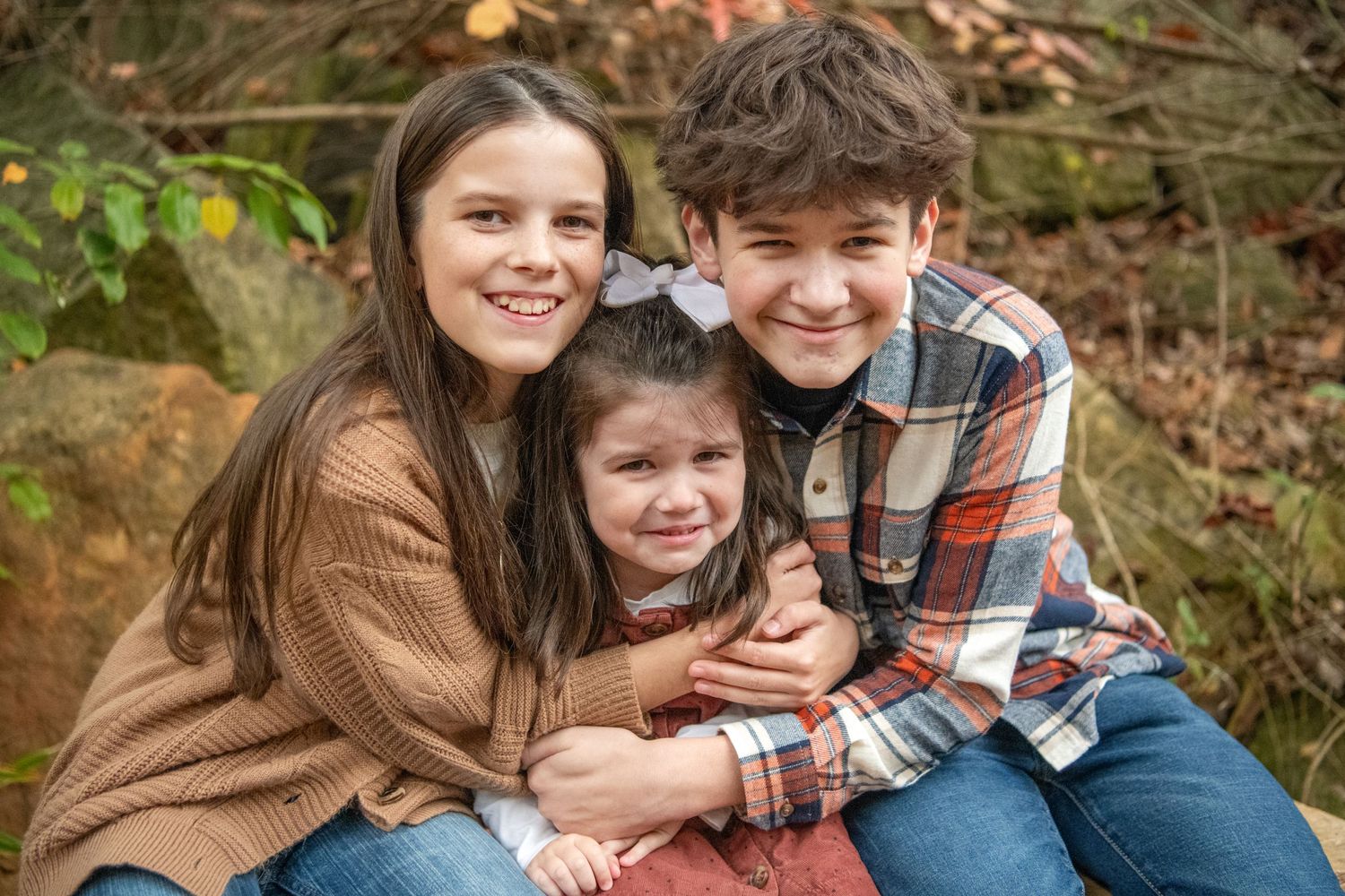 Three people embracing and smiling outdoors with fall foliage in the background.