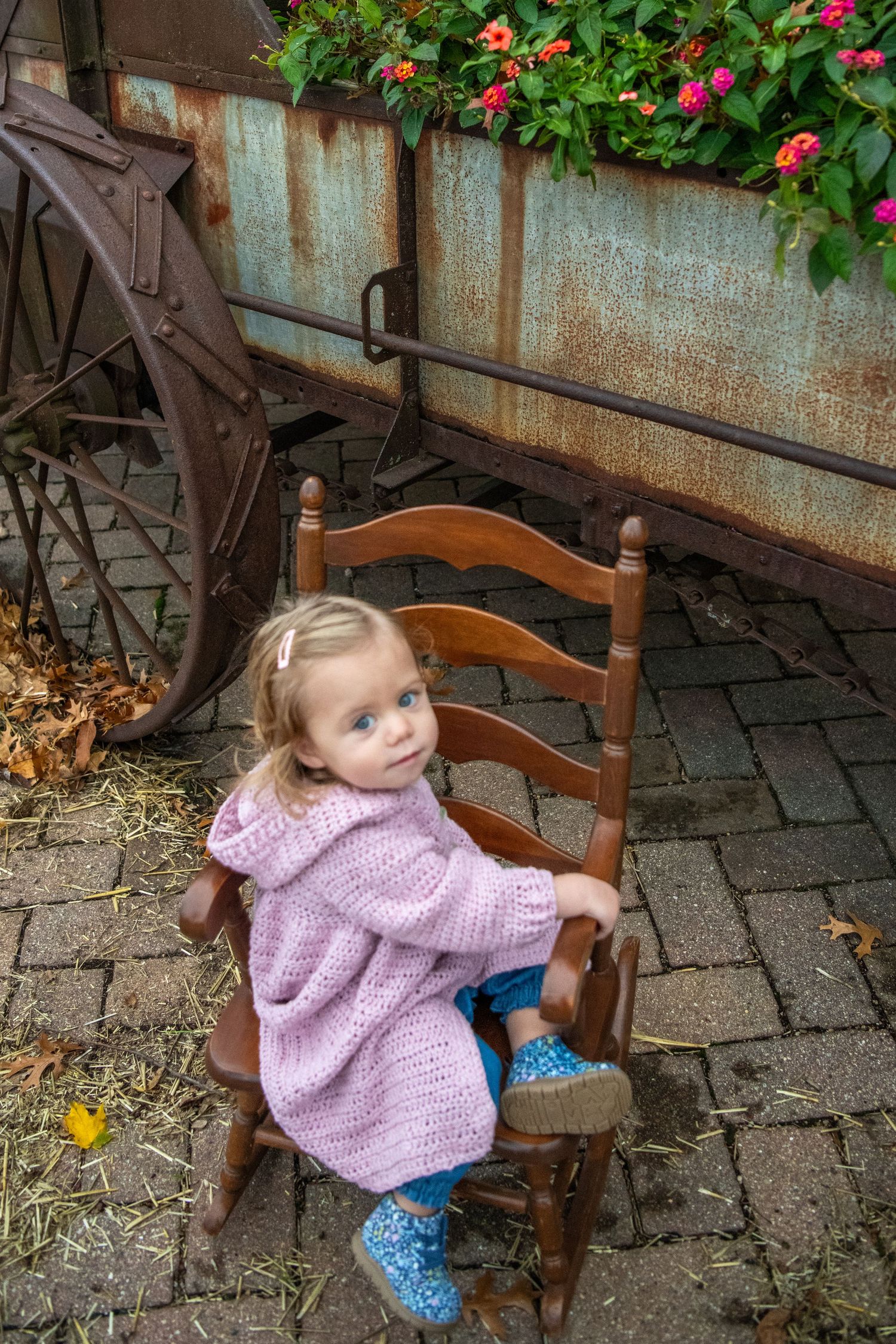 Child sitting in wooden rocking chair near rustic wagon wheel and colorful flowers.