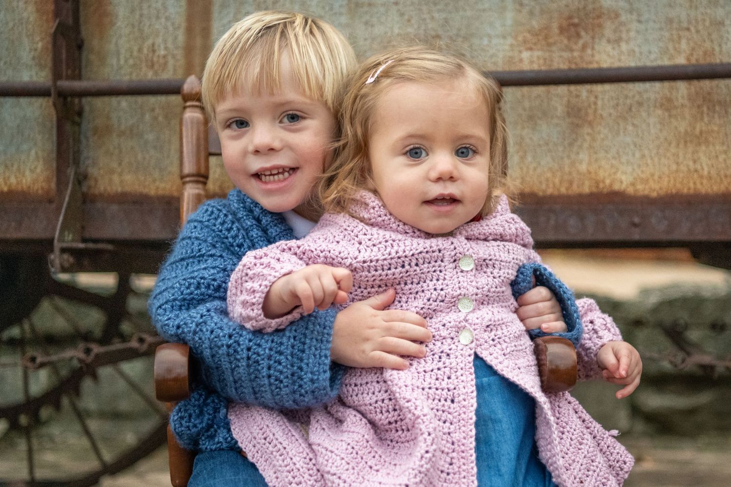 Two young children wearing knit sweaters sit together outdoors.