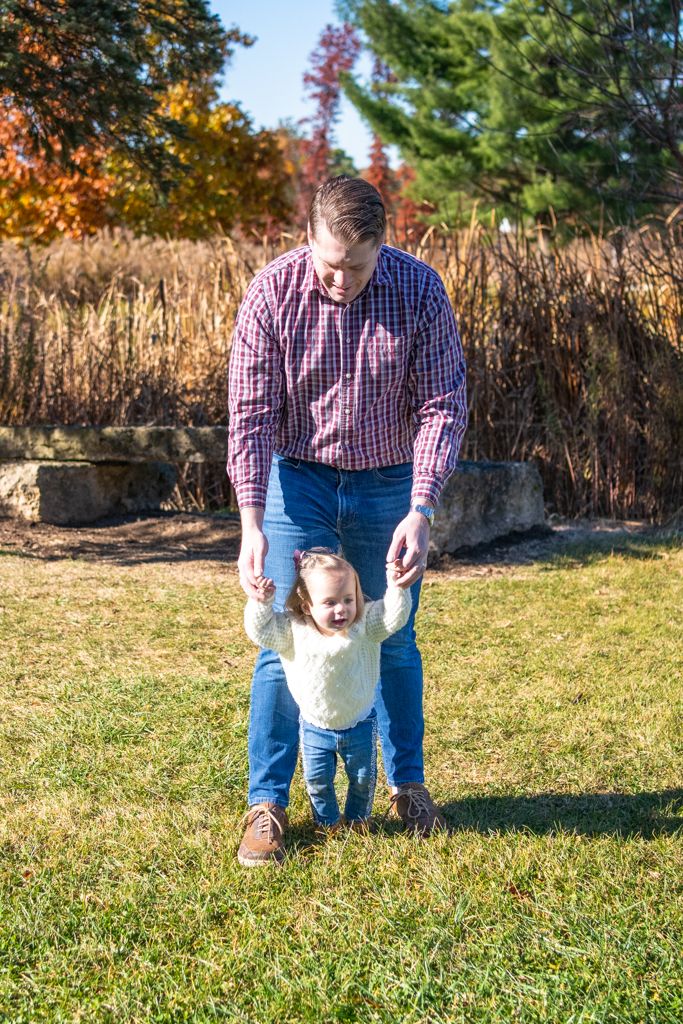 A parent helps a small child learn to walk on grass during autumn.