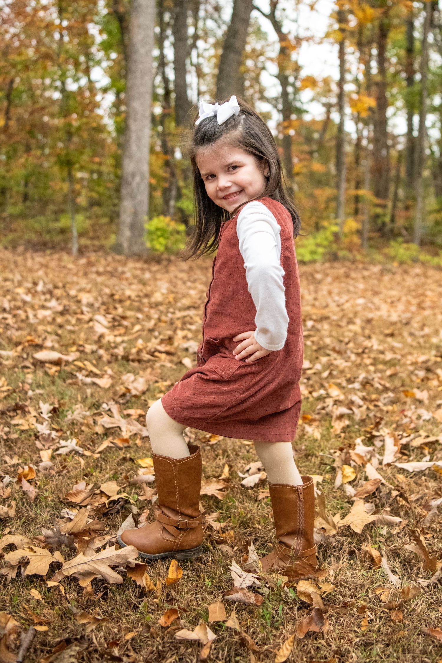 Young person in maroon dress and boots posing playfully among fallen autumn leaves.