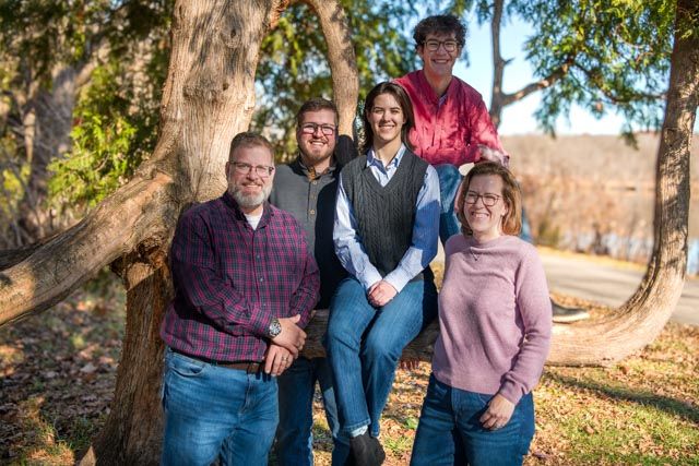 A group of people pose together outdoors against a backdrop of autumn trees and golden sunlight.
