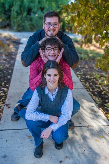 A couple poses playfully together outdoors on a sidewalk surrounded by autumn leaves and trees.