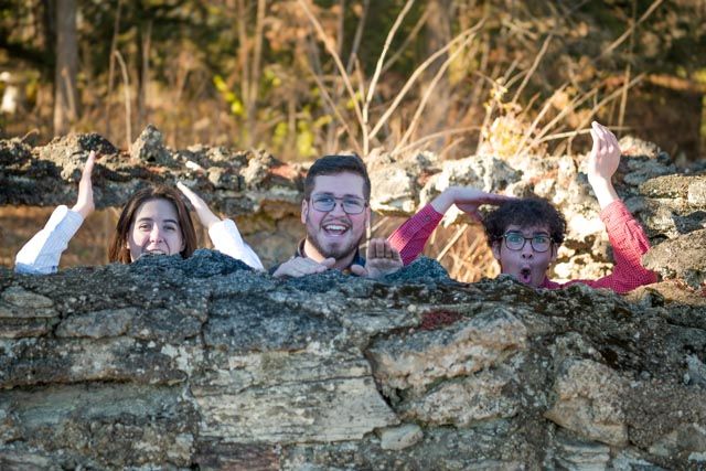 Group of people lying on ground and peeking over a dirt ledge while smiling at the camera.