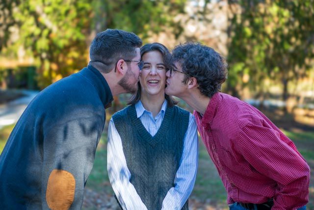A candid group photo shows three people laughing together in a park setting with trees in the background.