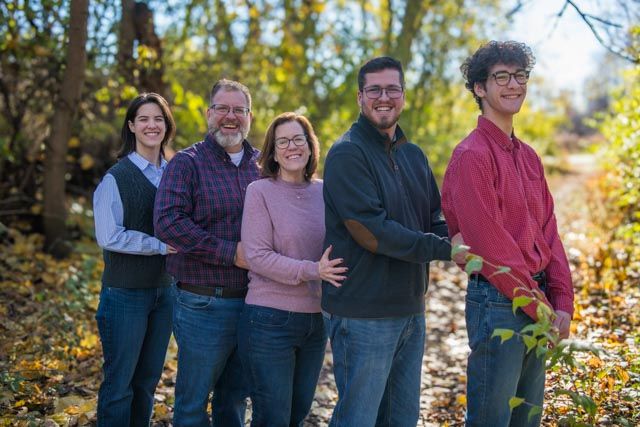 A group of five people stand together smiling outdoors on an autumn day with colorful fall foliage in the background.