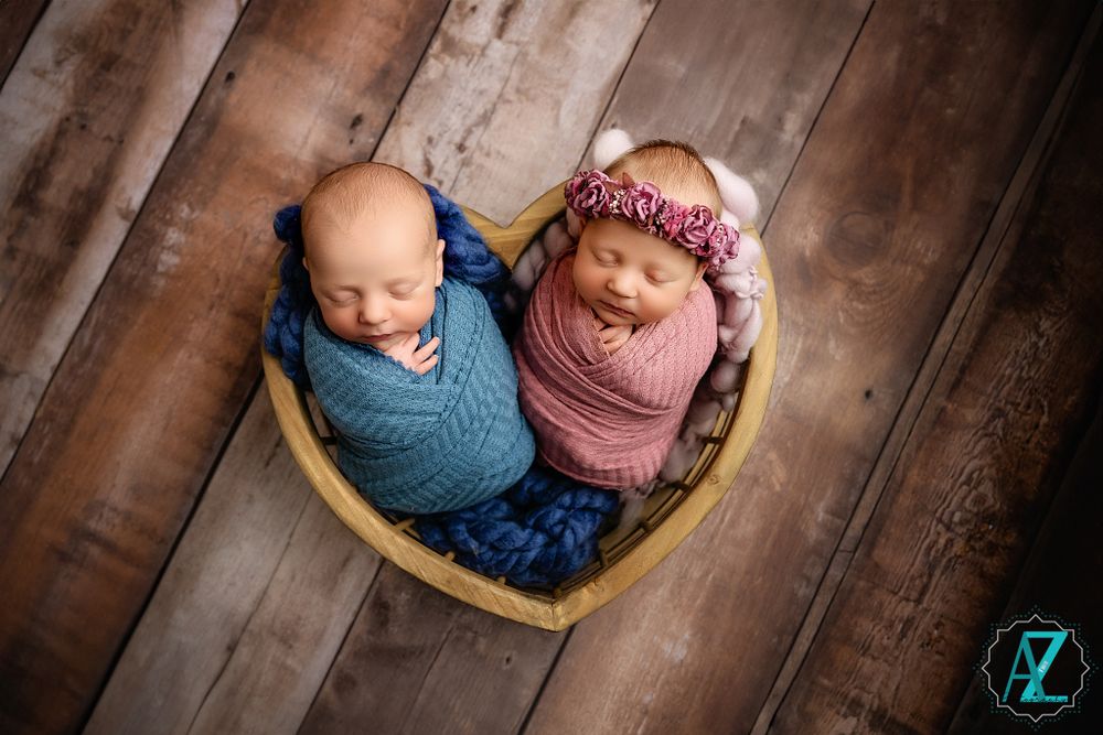 Two swaddled newborns nestled in a heart-shaped wooden bowl on rustic wooden floor.