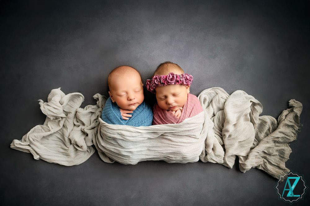 Two swaddled newborns rest together in soft white fabric against a dark background during professional photoshoot.