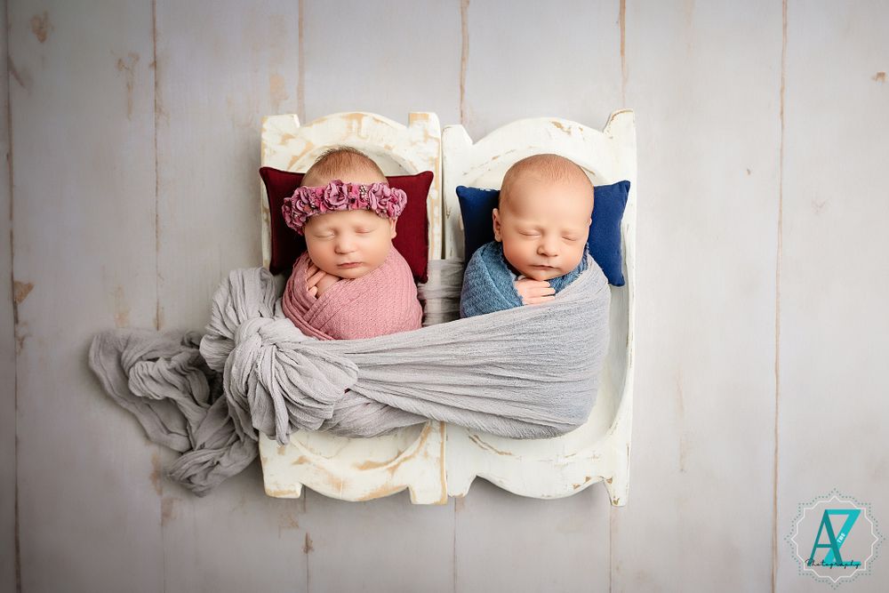 Two sleeping newborns swaddled in pink and blue wraps positioned side by side against white backdrop.