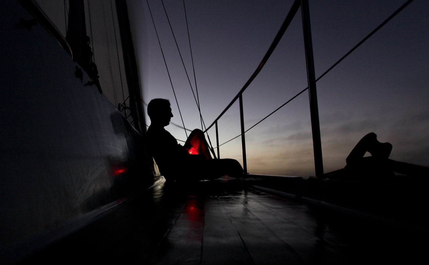 Silhouette figure sits on ship deck at sunset creating moody maritime scene.