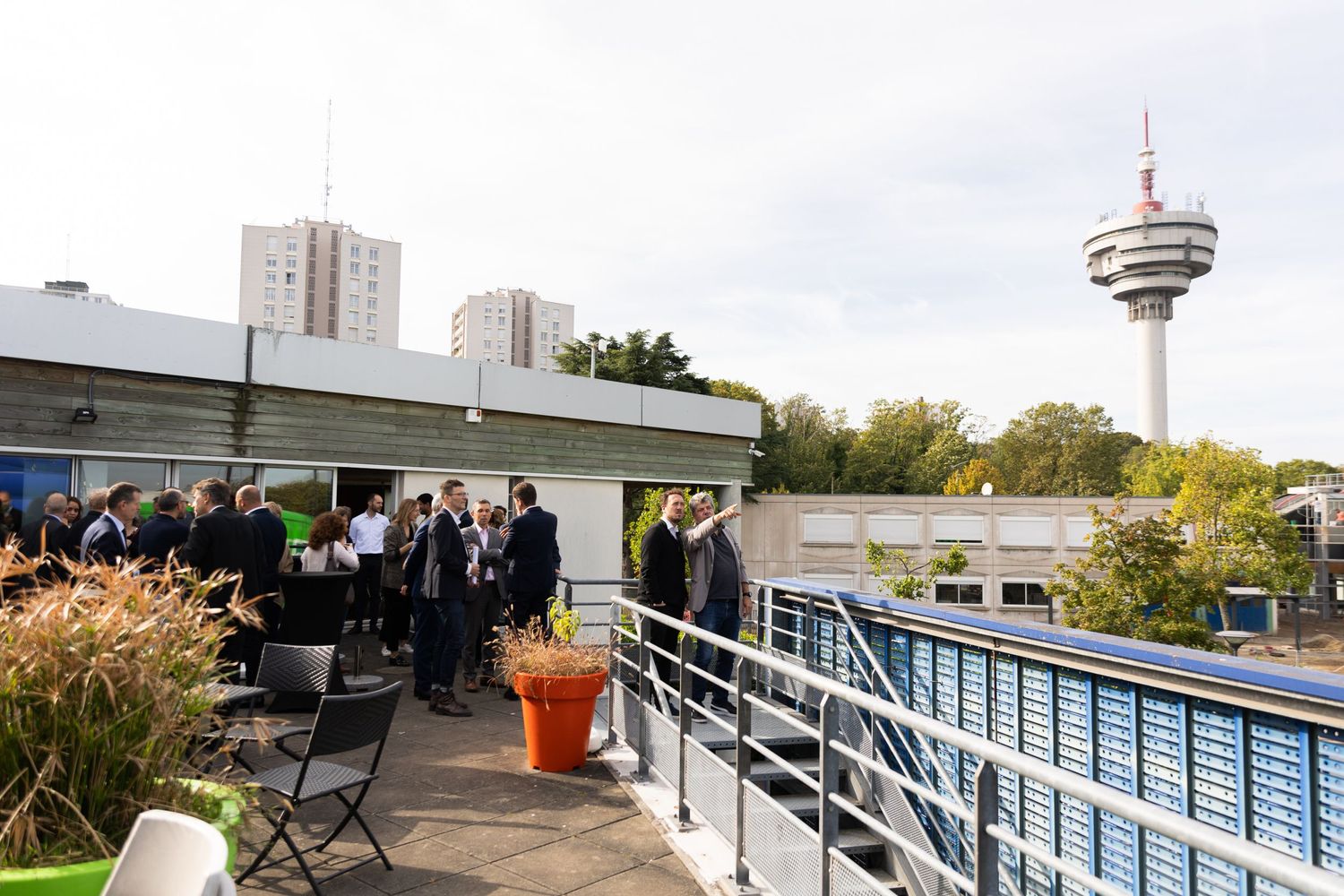People gather on a rooftop terrace with city views and a tall observation tower in the background on a cloudy day.