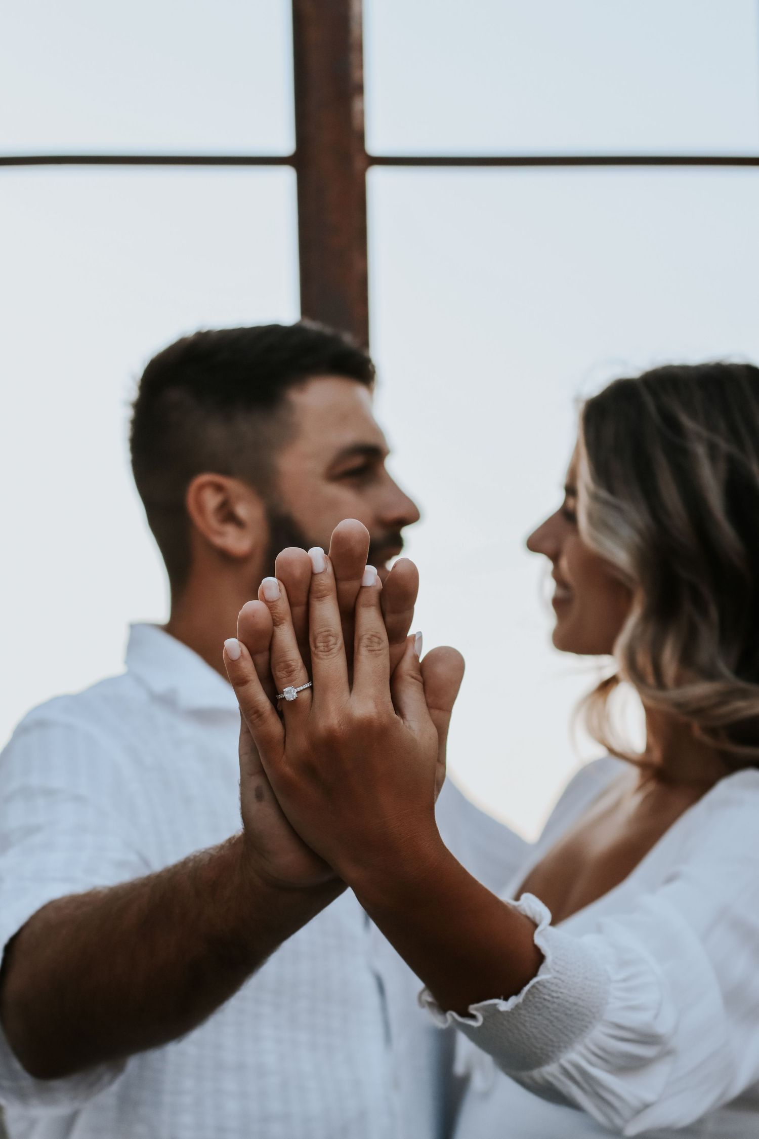 Couple in white outfits pressing hands together showing engagement ring, sharing a romantic moment indoors.