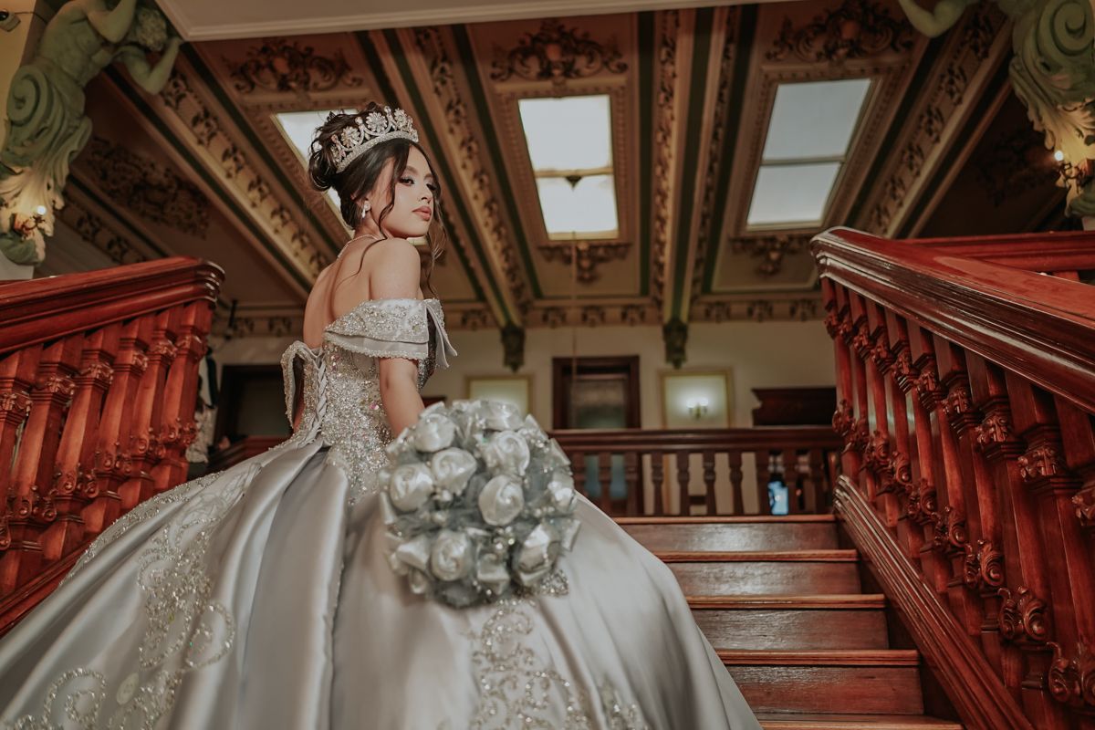 Elegant bride in embellished off-shoulder ballgown holds white bouquet while posing on ornate wooden staircase.