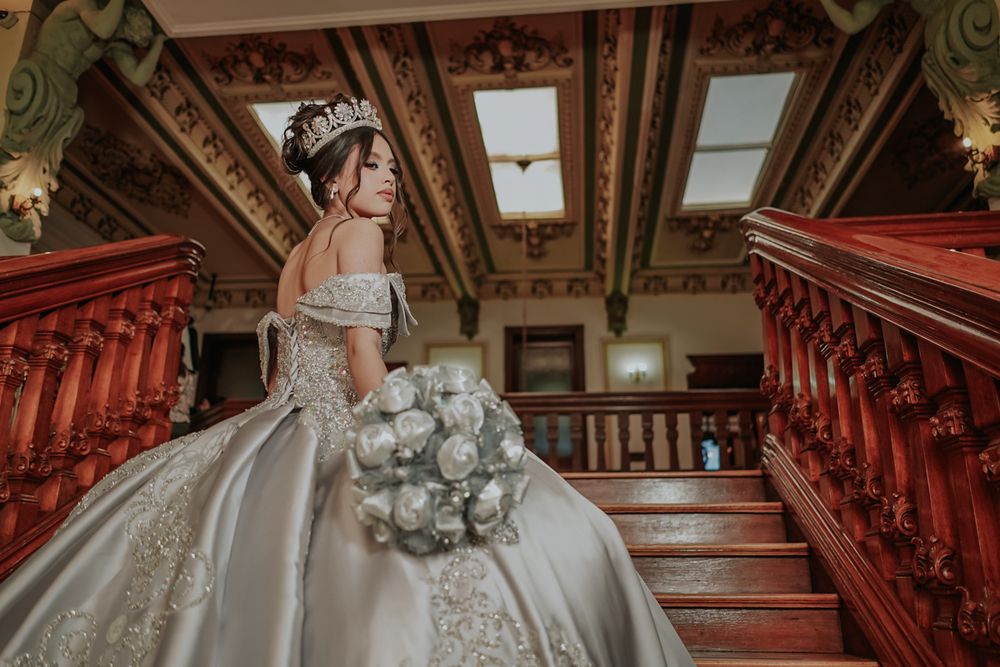 Elegant bride in embellished off-shoulder ballgown holds white bouquet while posing on ornate wooden staircase.