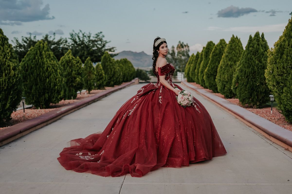A woman in a flowing burgundy ballgown poses elegantly along a stone pathway surrounded by green trees.