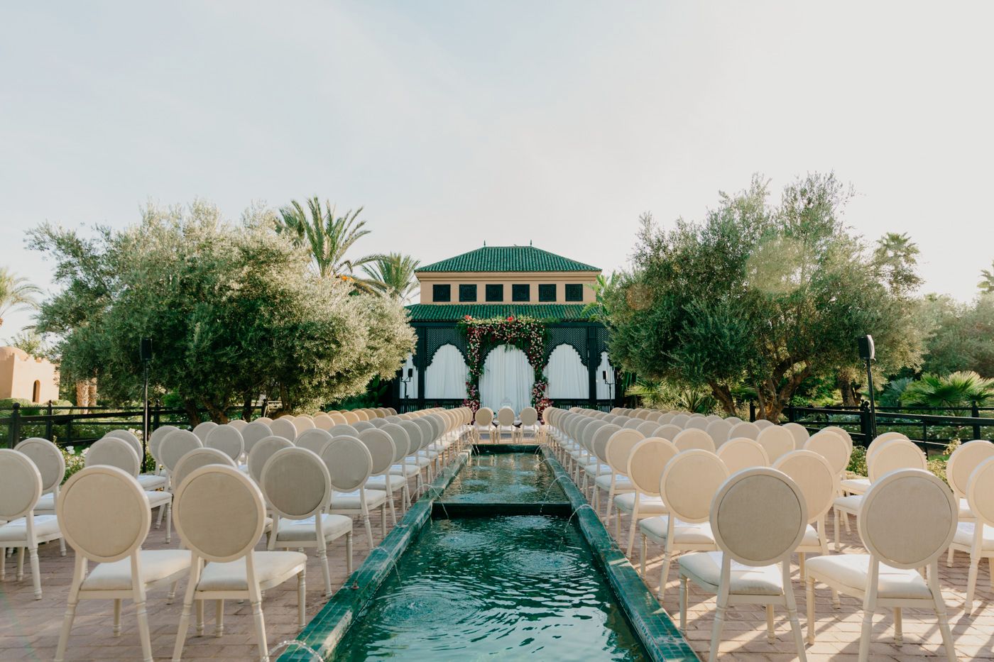Outdoor wedding ceremony setup with white chairs and water feature leading to a green gazebo.