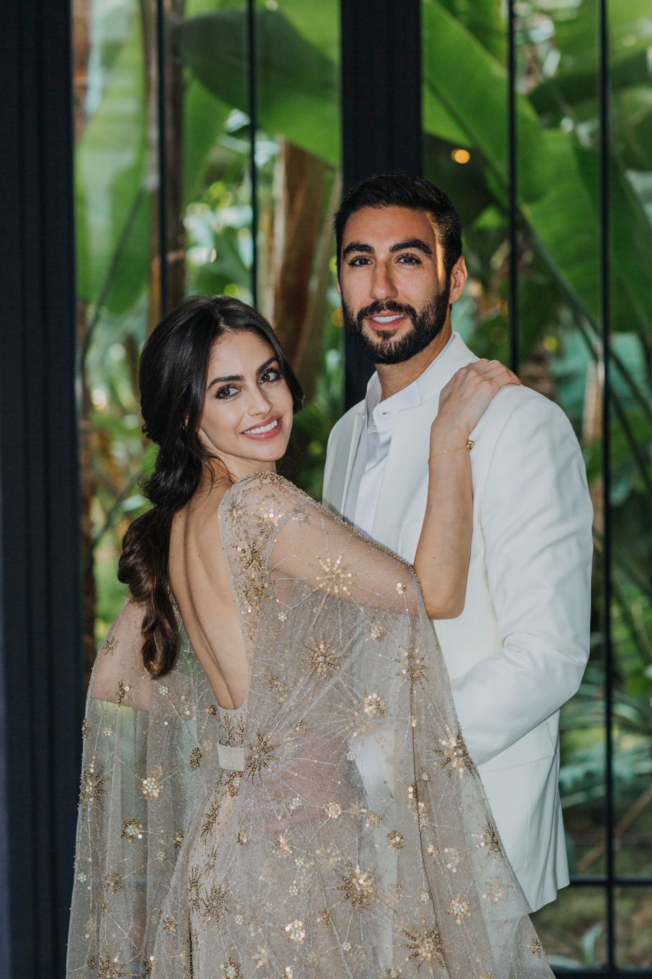 Couple in formal attire posing against tropical greenery backdrop.