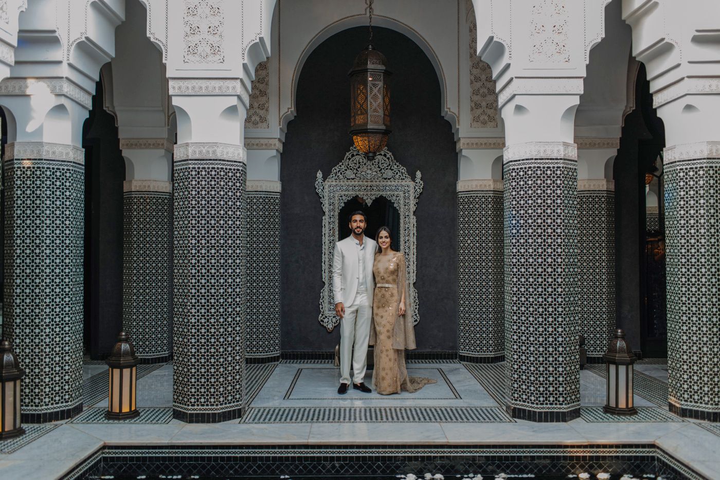 Couple posing in traditional attire under ornate Moroccan archway with geometric tile patterns and columns.