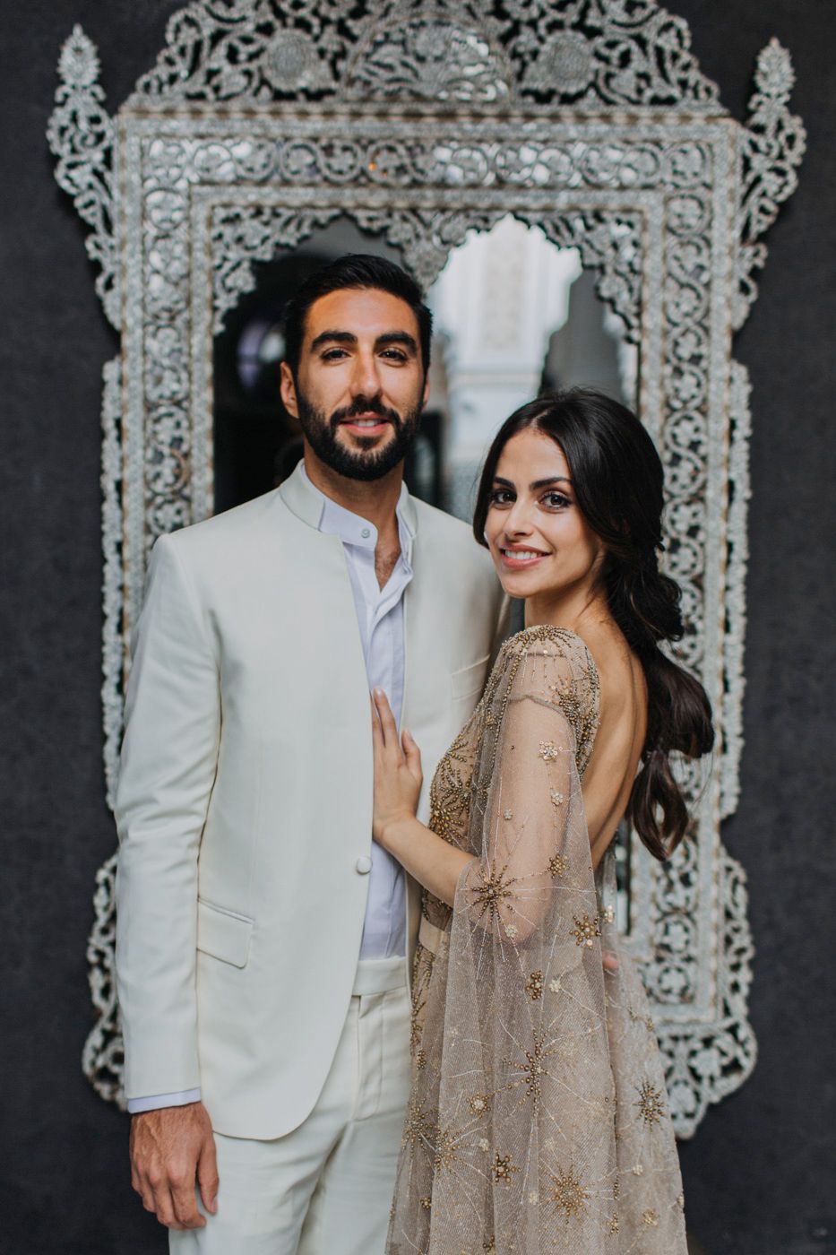 Stylish couple posing against ornate silver mirror frame in formal wedding attire.