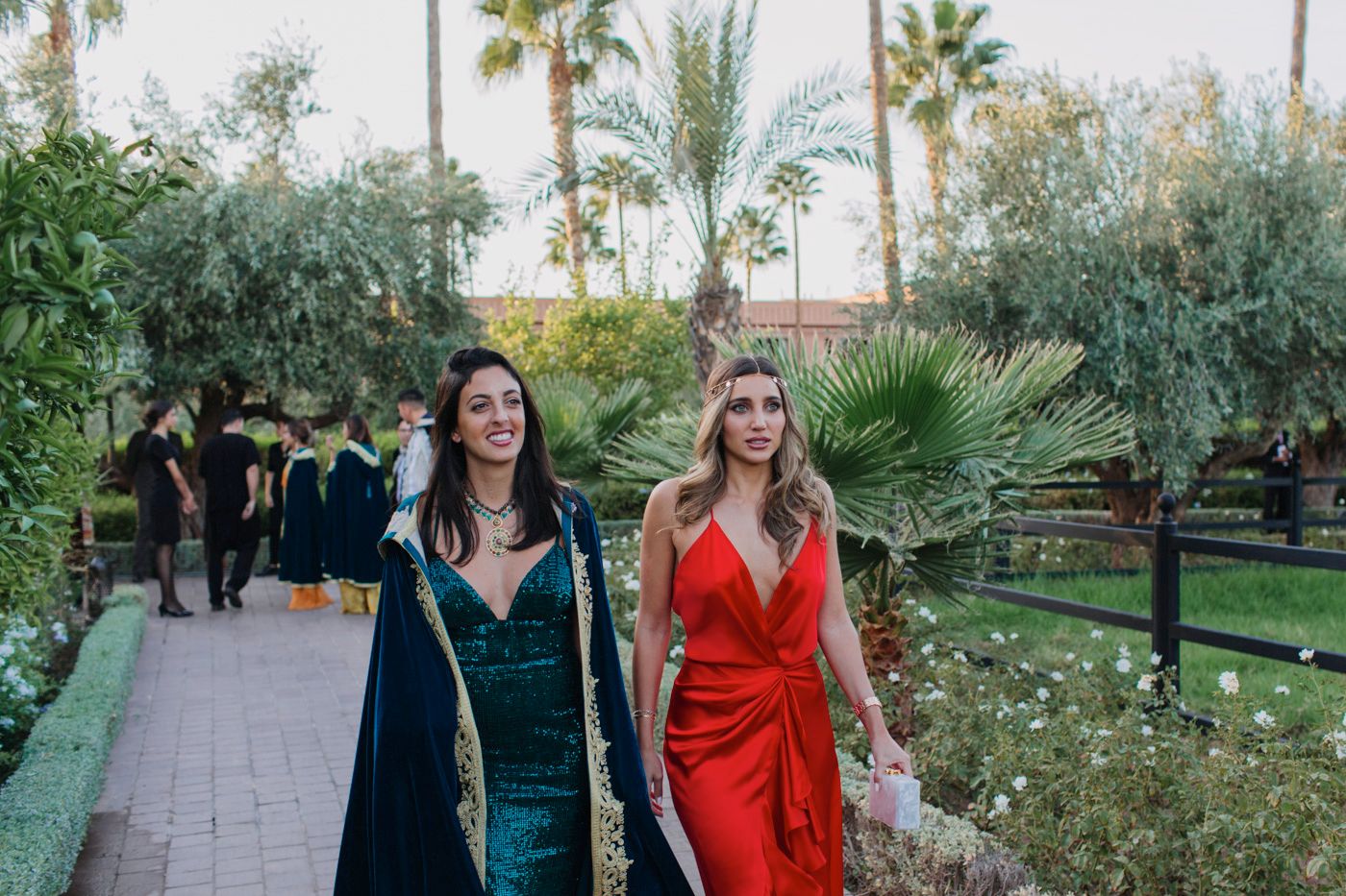 Two women walking through garden path wearing elegant evening gowns in jewel tones.