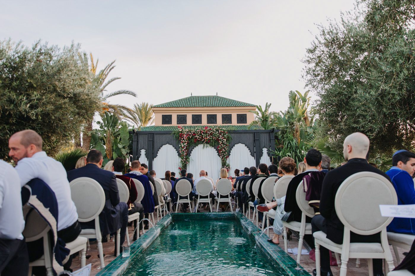Wedding ceremony setup with white chairs along pool reflecting Moroccan architecture.