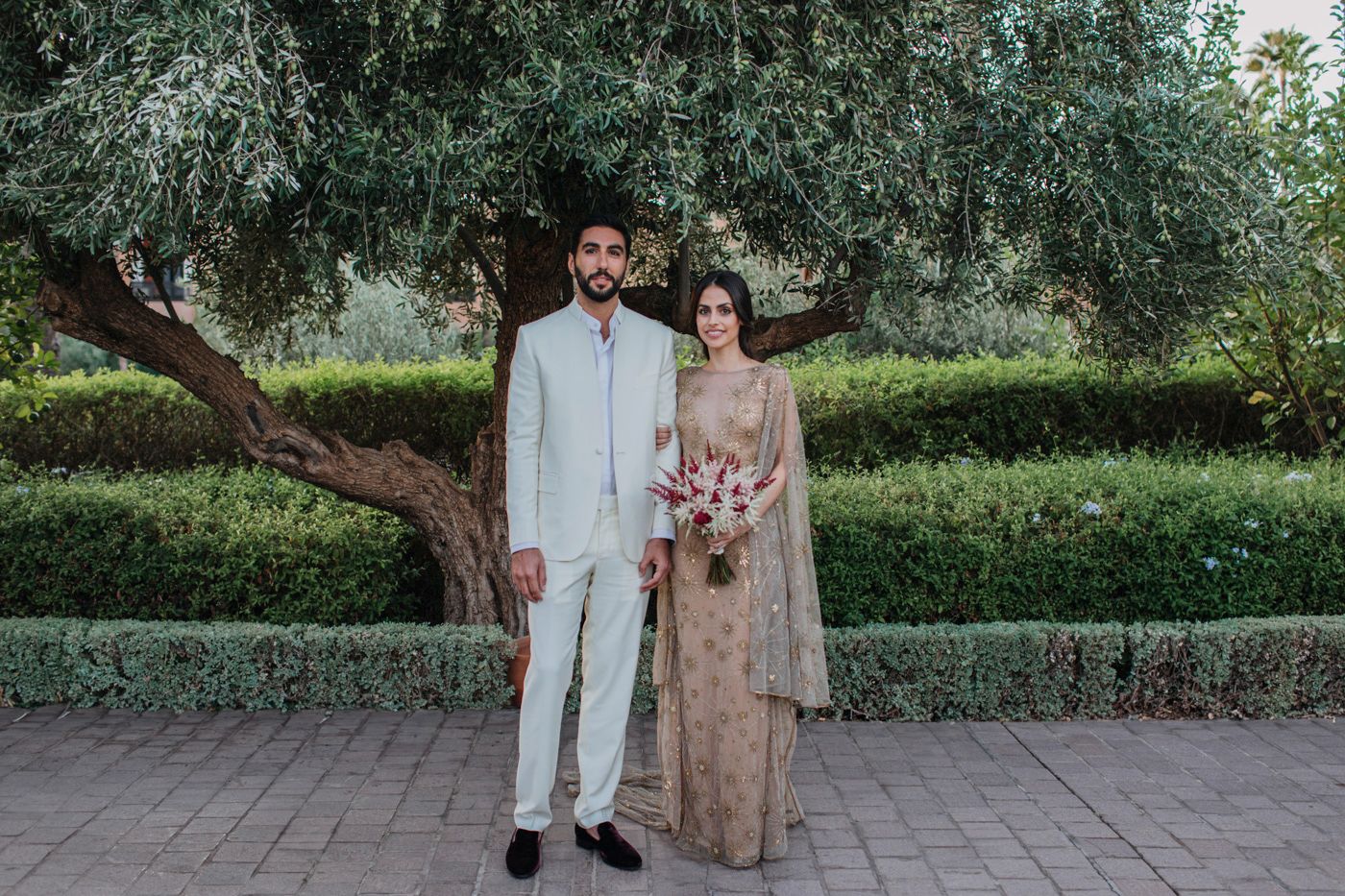 Stylish couple stands together in garden setting with white suit and embellished beige dress.