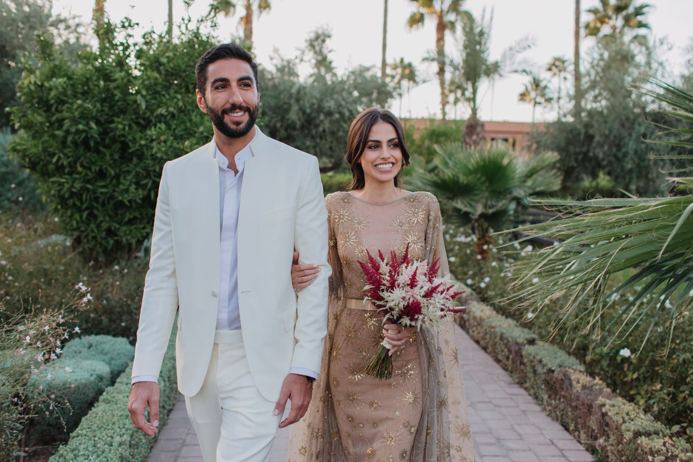Garden photoshoot with couple in white suit and beige dress holding dried flower bouquet.