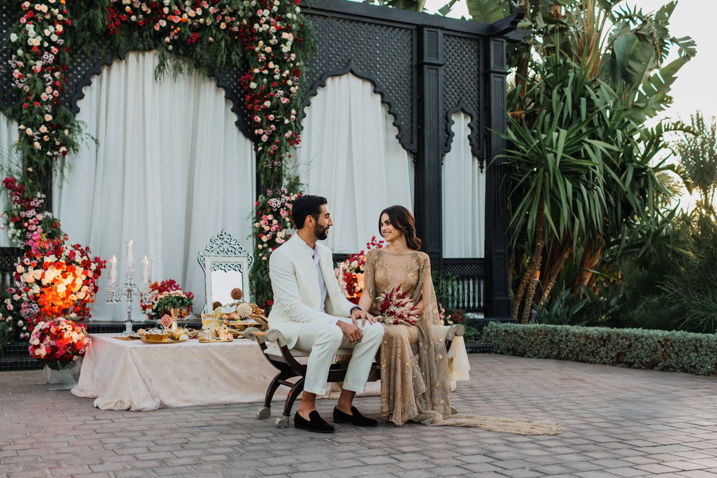 Romantic outdoor setting with floral arch backdrop and couple seated at decorated table.