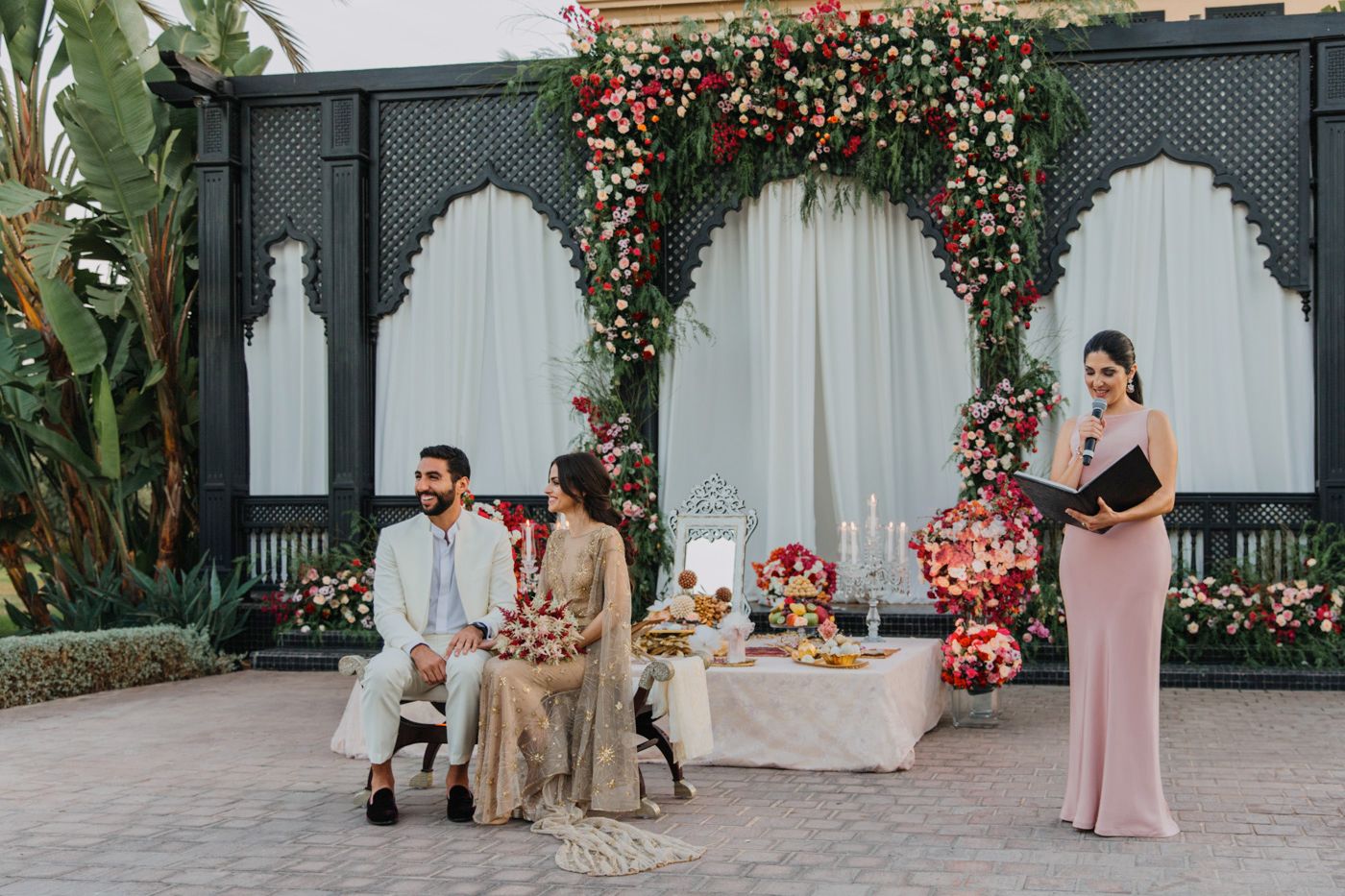 Outdoor celebration setup with decorative floral arch and seated couple at elegant table.
