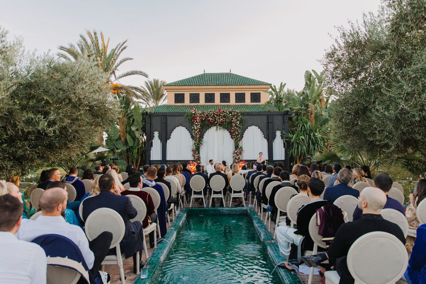 Wedding ceremony setup with chairs along pool leading to gazebo under palm trees.