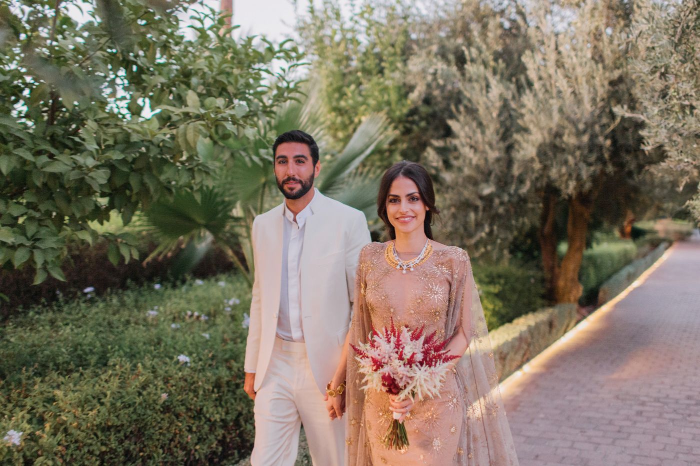 A couple in elegant attire walks along a garden path at dusk during what appears to be a wedding celebration.