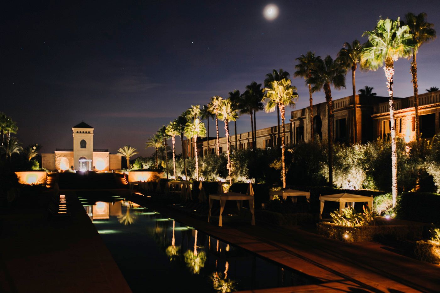 Illuminated palm trees line a reflective pool at night with Mediterranean-style architecture in the background.